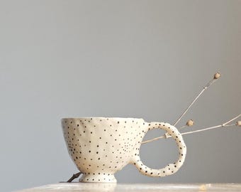 May include: A white ceramic mug with black polka dots and a unique handle. The mug is sitting on a light-colored surface.