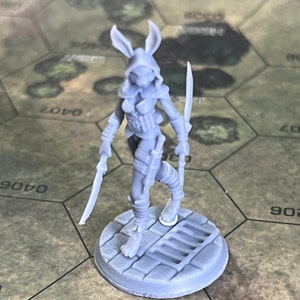 May include: A gray, 3D printed miniature of a female rabbit-like creature wearing a hooded cloak and holding two swords. The miniature is standing on a round base with a textured surface.