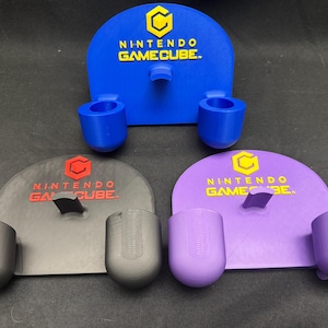 Peut inclure: Trois supports de manette Nintendo GameCube imprimés en 3D, bleus, gris et violets. Chaque support présente le logo et le texte GameCube. Le support bleu a deux supports de manette, tandis que les supports gris et violets en ont chacun deux.