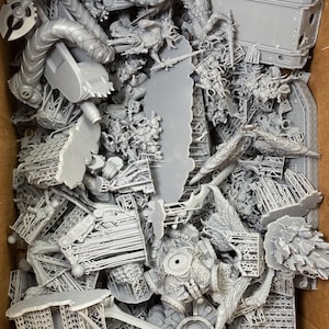 May include: A cardboard box overflowing with unassembled grey plastic miniatures. The collection includes figures, structures, and mechanical parts, indicating a tabletop gaming or model-building hobby.