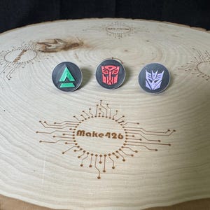 May include: Three round black and white buttons with different symbols. The buttons are on a wooden surface with a circuit board design and the text "Make426".