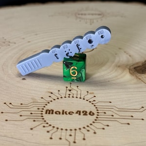 May include: A gray plastic tool with a series of numbered holes, used for sizing jewelry. The tool is lying on a wooden surface with a green six-sided die with gold numbers.