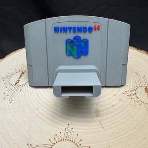 May include: A gray 3D-printed Nintendo 64 cartridge with the logo in blue, red, and green. The cartridge has a rectangular shape with a slot at the bottom. The text "NINTENDO 64" is visible on the front.