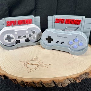 May include: Two grey Super Nintendo controllers on a wooden stand. The controllers are both grey and have the words "SUPER NINTENDO" printed on them. The stand is made of wood and has a circular shape. The stand has the words "Make 426" engraved on it.
