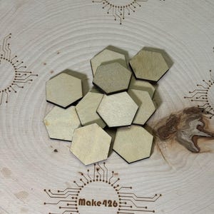 May include: A pile of eight unfinished wooden hexagonal game pieces on a light wood surface with a brown logo that says "Make426" in the center of the image.