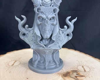May include: A gray bust of a skull-faced figure with large horns and ornate detailing. The sculpture has a detailed crown and a flowing cloak. The bust is on a round base, sitting on a wooden surface. The text "Make426" is visible.