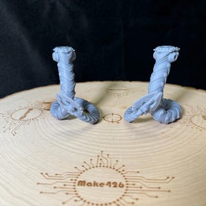 May include: Two gray 3D printed figures of a creature with a segmented body and a crown on its head. The figures are posed with their tails curled around their bodies. The figures are sitting on a wooden surface with a circular design.