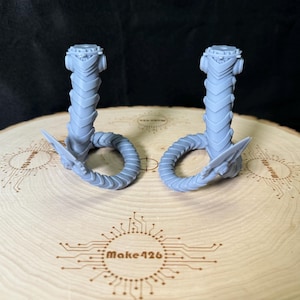 May include: Two light gray, 3D-printed cobra figurines. Each cobra has a detailed head, segmented body, and a curled tail. They are displayed on a wooden surface with the text "Make426" etched into it.
