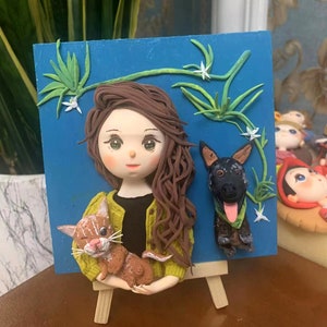 May include: A blue background with a hand-crafted clay figurine of a woman holding a cat. The woman has long brown hair and is wearing a green sweater. A black dog figurine is to the right of the woman. The background has green vines with white flowers.