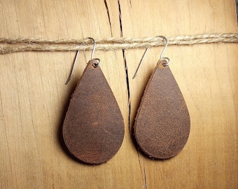 Peut inclure: Une paire de boucles d'oreilles en forme de goutte d'eau en cuir marron avec des crochets argentés suspendus à un morceau de bois.