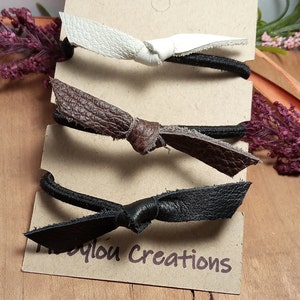 Puede incluir: Tres gomas para el pelo de cuero con un diseño de lazo anudado. Las gomas para el pelo son blancas, marrones y negras. Las gomas para el pelo se muestran en una tarjeta marrón con el texto "MyLou Creations".