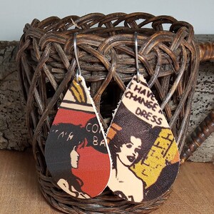 Può includere: Un paio di orecchini in pelle a forma di goccia con illustrazioni di ispirazione vintage. Un orecchino presenta una donna con il testo "Come Back" e l'altro orecchino presenta una donna con il testo "I Have Changed Dress".