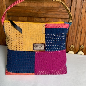 May include: A patchwork tote bag with a yellow zipper and a pink and yellow handle. The bag is made of fabric squares in yellow, navy blue, and fuchsia, with a stitched design. The label reads "साधना SEVA MANDIR".