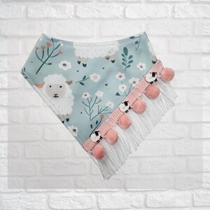 May include: A light blue bandana with a sheep and floral pattern. The bandana has a pink trim with pink pom-poms and white fringe. The sheep are white with black faces.