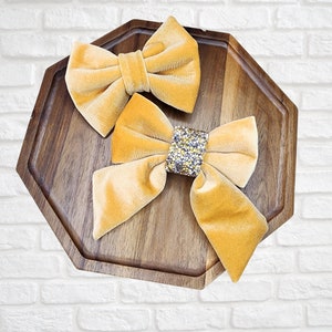 Puede incluir: Dos lazos de terciopelo de color amarillo mostaza. Un lazo tiene un gran adorno de pedrería en el centro.