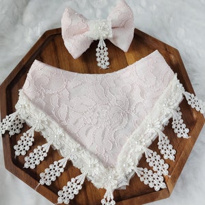 May include: A matching set of a pink lace bandana and bow tie with white lace trim and pearl accents. The bow tie has a pearl and flower center with a pearl drop. The set is displayed on a wooden tray.