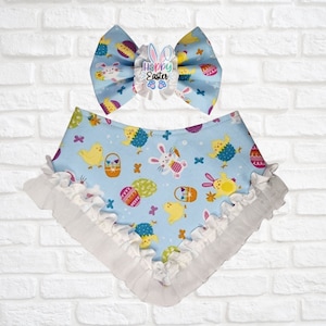 May include: A light blue Easter-themed baby bow and bandana set. The bow features a "Happy Easter" embellishment. The bandana and bow are decorated with Easter eggs, chicks, and bunnies. The bandana has a white ruffled edge.