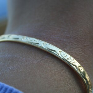 May include: A thin, polished gold-tone bangle bracelet featuring a repeating embossed pattern of small fish along the entire outer surface. The metal band has a smooth, reflective finish and a rounded profile, encircling the wrist with a consistent design.