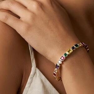 May include: Gold bracelet adorned with a variety of colourful gemstones in different shapes. The gemstones include red, purple, green, yellow, and blue. The bracelet is worn on a wrist, with a person's arm and shoulder visible.