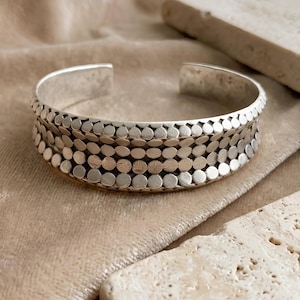 May include: A silver cuff bracelet with a textured design of small, evenly spaced circles. The bracelet has an open design and rests on a beige fabric surface. The jewelry is a stylish accessory.