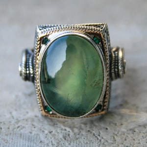 May include: A silver ring featuring a large, oval, green gemstone. The ring has a gold-coloured border around the gemstone and small green accent stones. The band has decorative detailing.