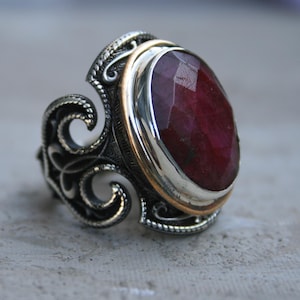 May include: A silver ring featuring a large, oval-shaped, faceted deep red gemstone set in a gold-toned bezel. The band is crafted with intricate, dark-oxidised silver scrollwork and filigree patterns that extend along the sides of the ring.
