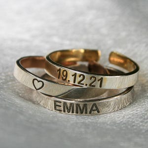 Peut inclure: Deux bagues en argent avec du texte gravé. La bague du haut porte la date "19.12.21" gravée. La bague du bas porte le nom "EMMA" gravé. Les bagues sont empilées l'une sur l'autre.