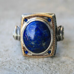 Puede incluir: Un anillo de plata y oro con una gran piedra ovalada de lapislázuli azul oscuro. El anillo tiene un engaste rectangular adornado con pequeñas piedras azules y detalles laterales decorativos. Un anillo llamativo.