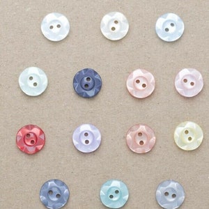May include: Assortment of 16 two-hole buttons in various colours, including white, pink, blue, red, and yellow. The buttons have a starburst design and are arranged in a grid pattern.