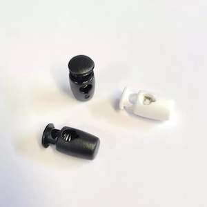 May include: Three cord locks are shown. Two are black, and one is white. These plastic cord locks have a cylindrical shape and a spring mechanism for tightening cords. They are used in various applications.