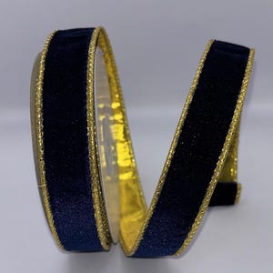 Puede incluir: Un rollo de cinta de terciopelo azul marino con un borde dorado brillante. La cinta está enrollada, mostrando las texturas y colores contrastantes. Ideal para manualidades, envolver regalos o decoraciones festivas.