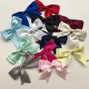 May include: A collection of 15 satin ribbon bows in various colours, including white, black, blue, red, pink, yellow, green, and grey. The bows are arranged in a random pattern on a white surface.