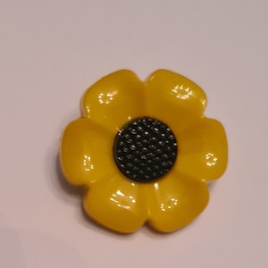 Może przedstawiać: Żółty guzik w kształcie kwiatu z czarnym środkiem. Guzik ma pięć płatków i błyszczące wykończenie.