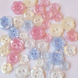 May include: Assortment of pastel coloured flower shaped buttons with two holes each. The buttons are in shades of pink, blue, and white.