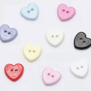 Könnte beinhalten: Sortiment von zehn herzförmigen Knöpfen in verschiedenen Farben, darunter Blau, Lila, Weiß, Gelb, Rot, Rosa und Schwarz. Jeder Knopf hat zwei Löcher.