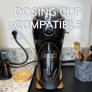 May include: A black Niche Zero coffee grinder with a stainless steel dosing cup attached to the bottom. The text "DOSING CUP COMPATIBLE" is written in white letters on the grinder.