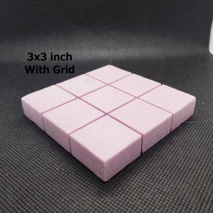 May include: A light pink, square foam grid, approximately 7.6 x 7.6 cm, with nine individual squares. The text "3x3 inch With Grid" is visible in the upper left corner. The grid is on a dark, textured surface.
