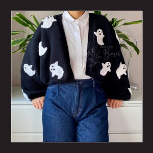 May include: A black cropped cardigan with white ghost appliques. The cardigan is knitted and has a relaxed fit. The text "knits by Basil" is visible on the right side of the cardigan.