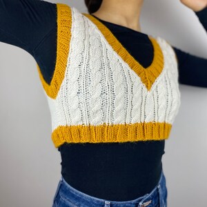 May include: A white and mustard yellow cable-knit sweater vest. The vest has a V-neck and a ribbed hem. The cable knit pattern is visible throughout the vest. The vest is worn over a black long-sleeve shirt and blue jeans.