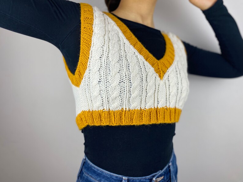 May include: A white and mustard yellow cable-knit sweater vest. The vest has a V-neck and a ribbed hem. The cable knit pattern is visible throughout the vest. The vest is worn over a black long-sleeve shirt and blue jeans.
