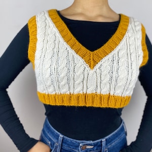 May include: A cropped cable-knit vest in cream and mustard yellow. The vest features a V-neck and a ribbed hem. The cream-colored front panel has a cable knit design, and the edges are trimmed with mustard yellow.