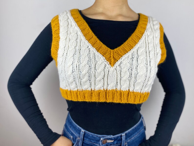 May include: A cropped cable-knit vest in cream and mustard yellow. The vest features a V-neck and a ribbed hem. The cream-colored front panel has a cable knit design, and the edges are trimmed with mustard yellow.