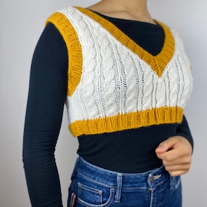 May include: A white cable-knit vest with a mustard yellow trim and V-neck. The cropped sweater has a classic design and is made of a soft, textured material. The vest is worn over a black long-sleeve shirt.