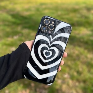 May include: Black phone case with a white swirling heart design. The case has a raised camera bump with three lenses.