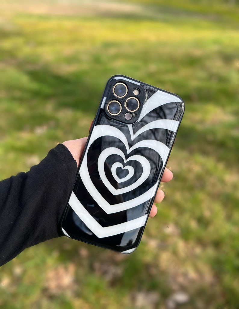 May include: Black phone case with a white swirling heart design. The case has a raised camera bump with three lenses.