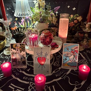 May include: A mystical scene featuring tarot cards, candles, and floral arrangements. The cards include "The Tower", "Three of Swords", and "Knight of Swords". Pink candles and a decorative lamp add to the ambiance. A black cloth with zodiac symbols is the backdrop.