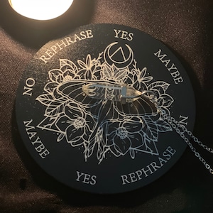Puede incluir: Un tablero de adivinación circular negro con un diseño de polilla blanca y las palabras "YES", "MAYBE", "NO" y "REPHRASE" alrededor del borde. Un péndulo de cristal transparente cuelga de una cadena plateada.