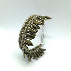 Könnte beinhalten: Ein handgefertigtes Armband mit olivgrünen Metallic-Akzenten. Das Armband besteht aus kleinen, perlenartigen Elementen und größeren, blattförmigen Teilen, die durch eine silberne Kette verbunden sind. Die Gesamtästhetik ist elegant und künstlerisch.