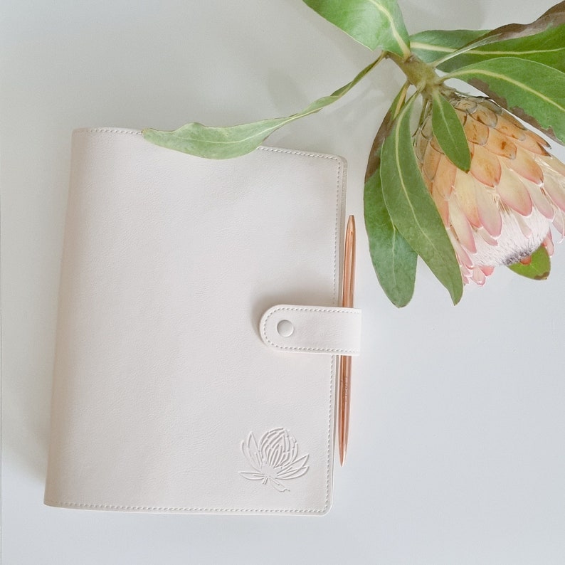 May include: A white leather planner with a gold pen and a floral design embossed on the front cover.