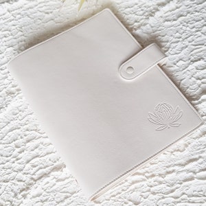 May include: A white leather planner with a floral design embossed on the front cover. The planner has a flap closure with a button.